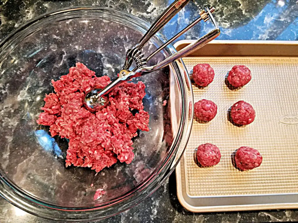 meatball mix in bowl with small cookie scoop next to cookie sheet with formed meatballs